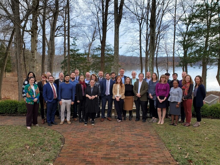 A group of scholars, practitioners, policymakers, and funders poses for a group photo against a backdrop of late winter at the Point of View Retreat and Research Center in Lorton, VA.